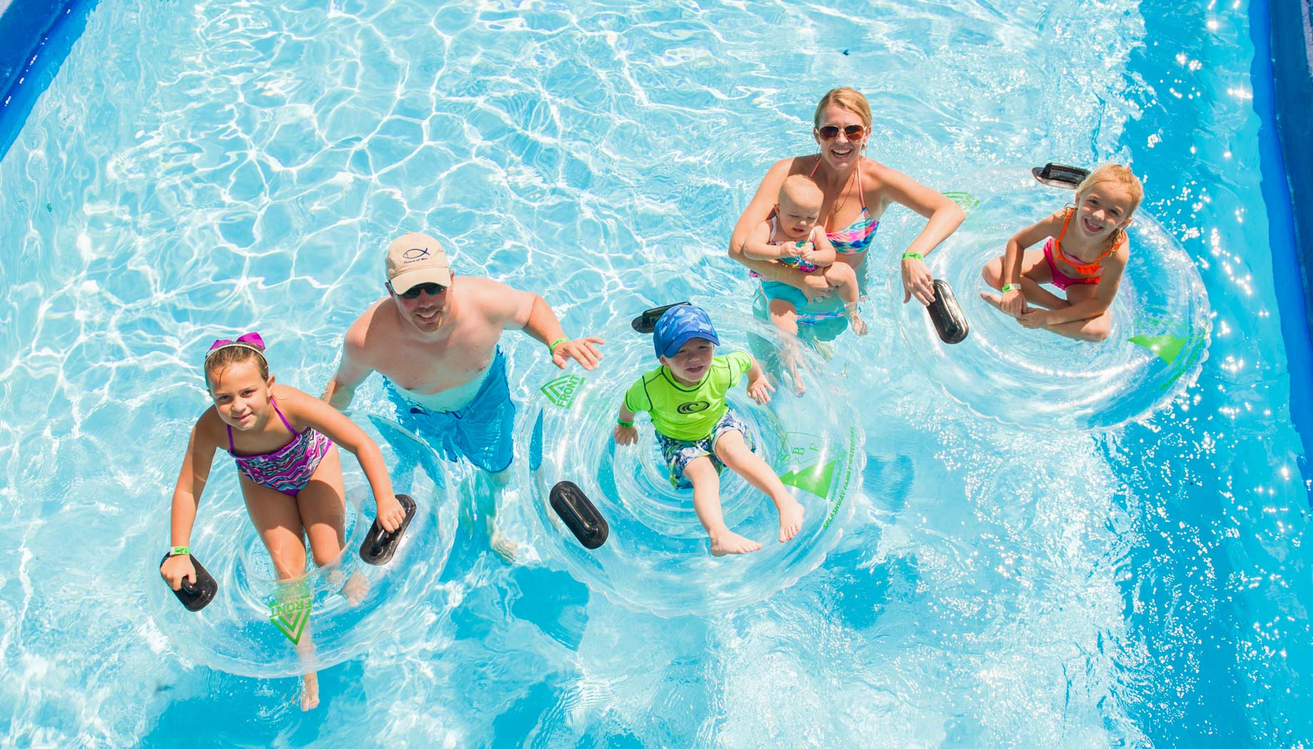 lazy-river-family A group of five people, two adults and three children, float in a lazy river using inflatable rings on a sunny day.