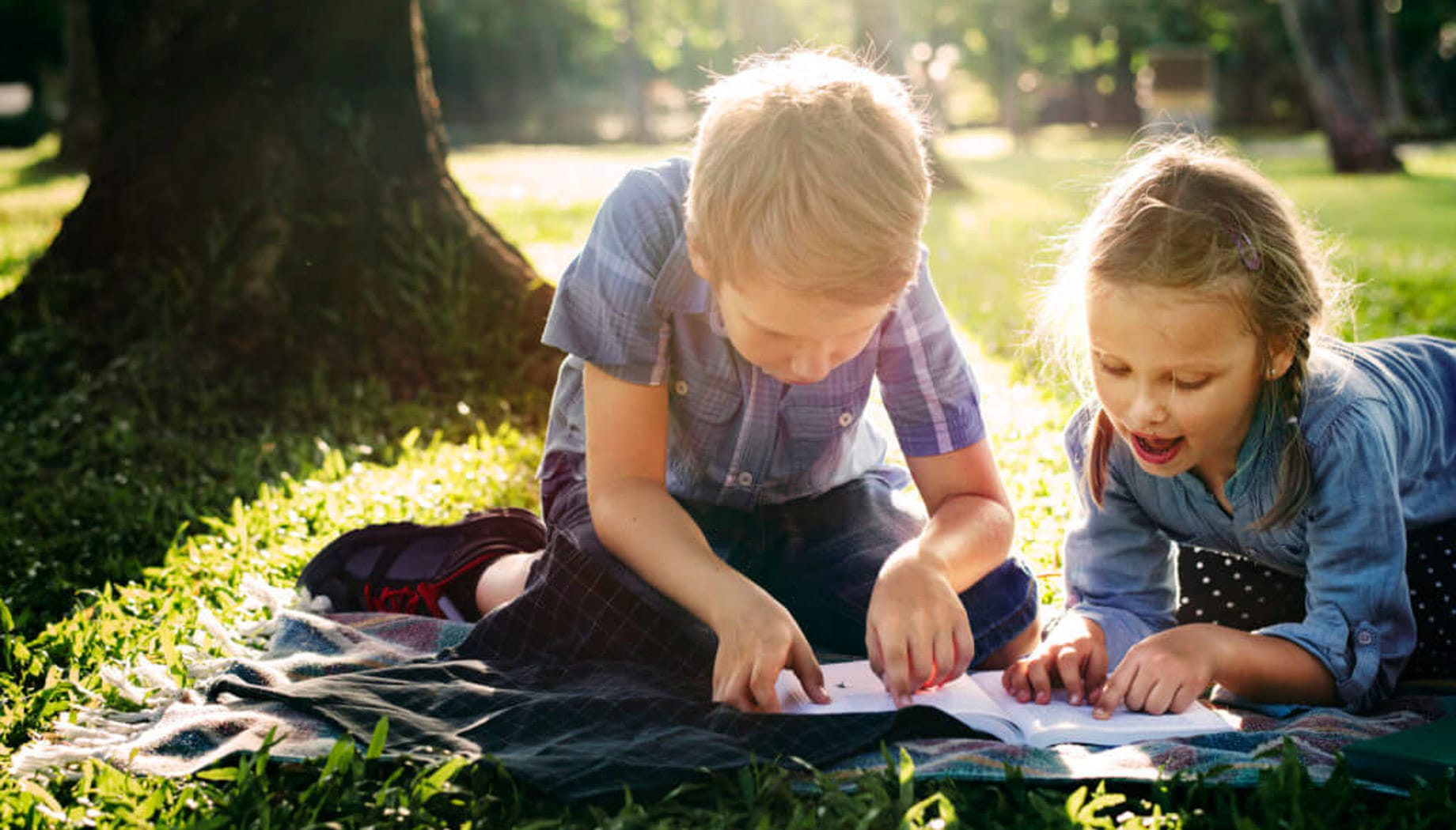 Two children lie on a blanket outside, focused on a book.
