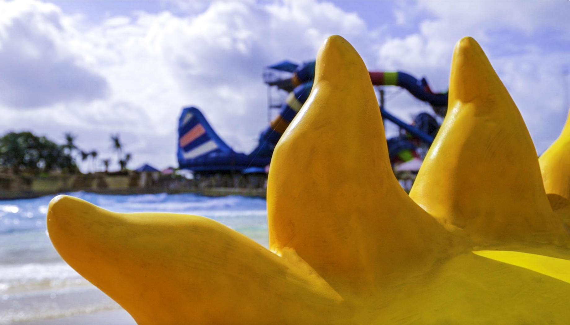 BLOG Close-up of a yellow sun-shaped sculpture with a water park featuring colorful slides in the background against a partly cloudy sky.