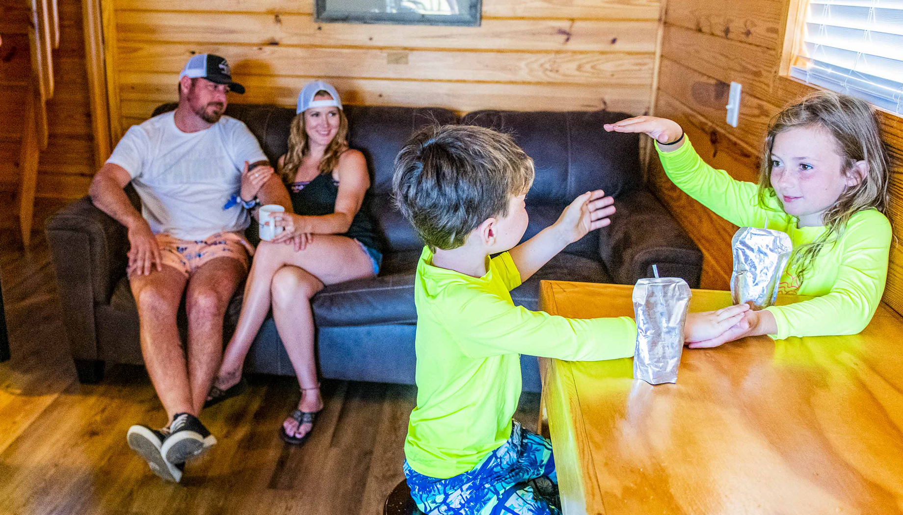 BLOG Two children in swimwear sit at a wooden table, playing and drinking from pouches. Behind them, two adults relax on a couch in a wood-paneled room.
